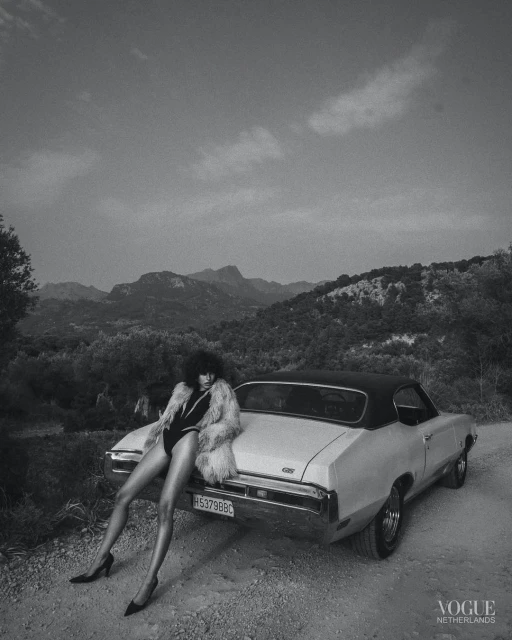 Black-and-white shot of a woman in a bodysuit and fur coat leaning against a vintage car in a mountainous rural setting.