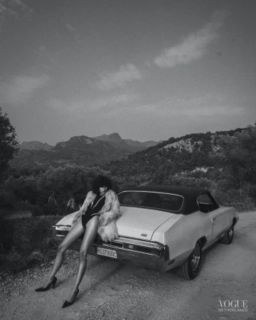 Black-and-white shot of a woman in a bodysuit and fur coat leaning against a vintage car in a mountainous rural setting.