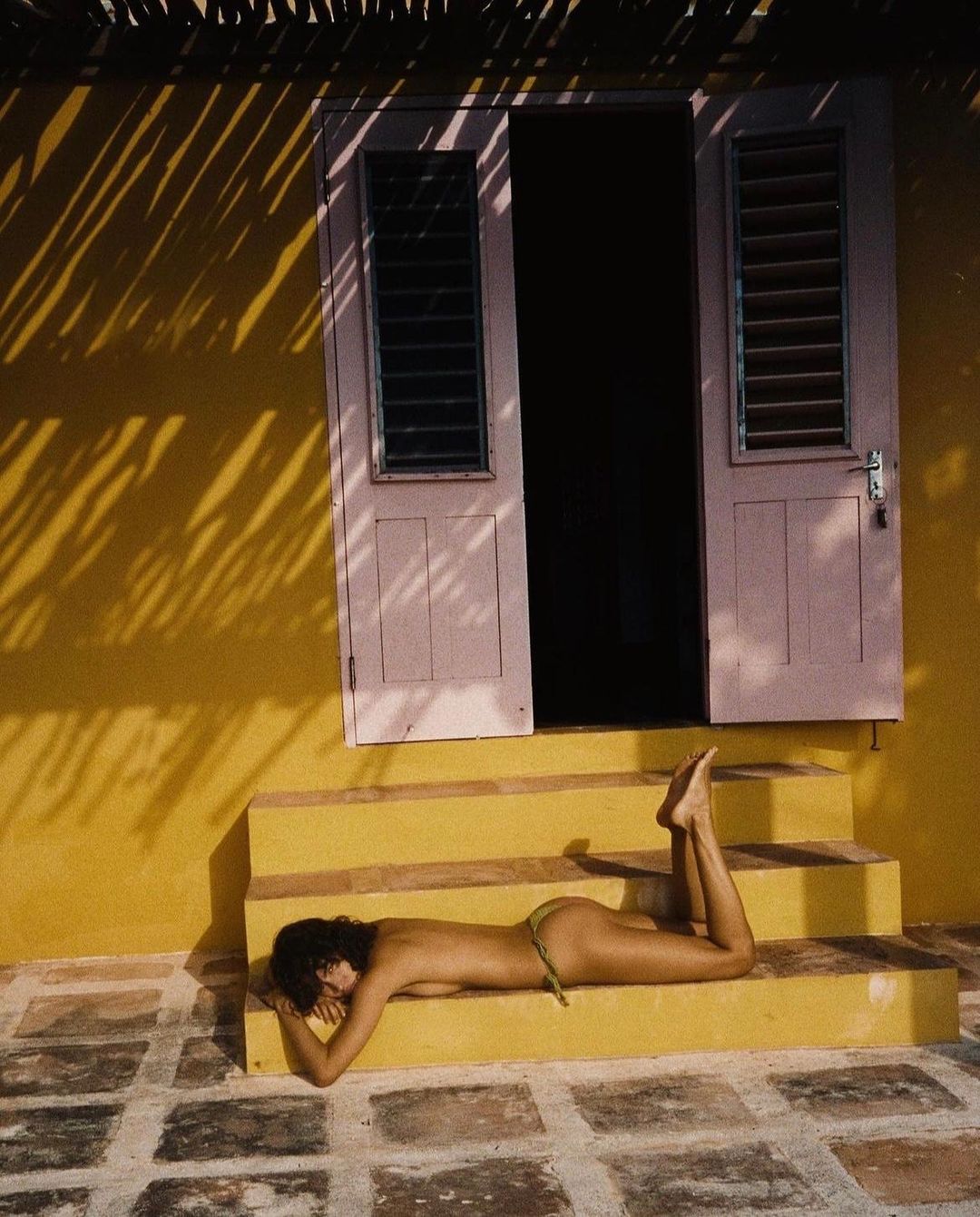 Topless woman sunbathes on yellow steps near a pink door with black shutters; shadows from palm fronds overlay the scene.