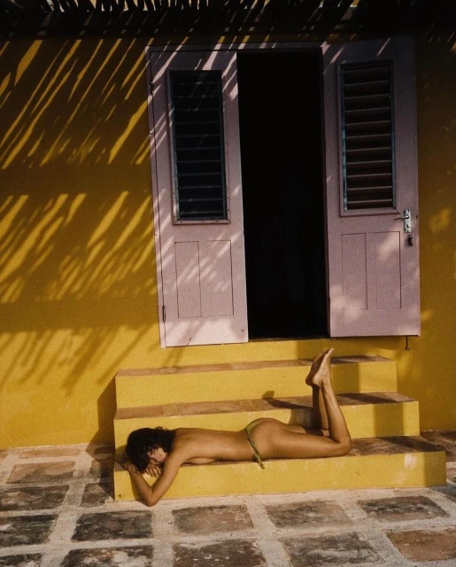 Topless woman sunbathes on yellow steps near a pink door with black shutters; shadows from palm fronds overlay the scene.