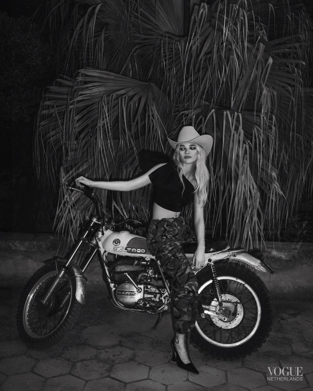 Blonde woman in cowboy hat poses with a vintage motorcycle against a palm tree backdrop in a black and white Vogue Netherlands editorial shot.