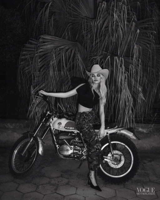 Blonde woman in cowboy hat poses with a vintage motorcycle against a palm tree backdrop in a black and white Vogue Netherlands editorial shot.