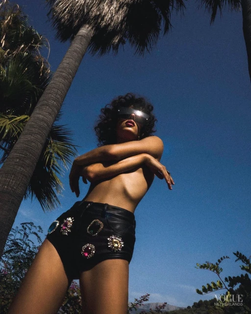 Adult female fashion model poses in black jeweled shorts against a clear blue sky backdrop with palm trees and lush greenery.