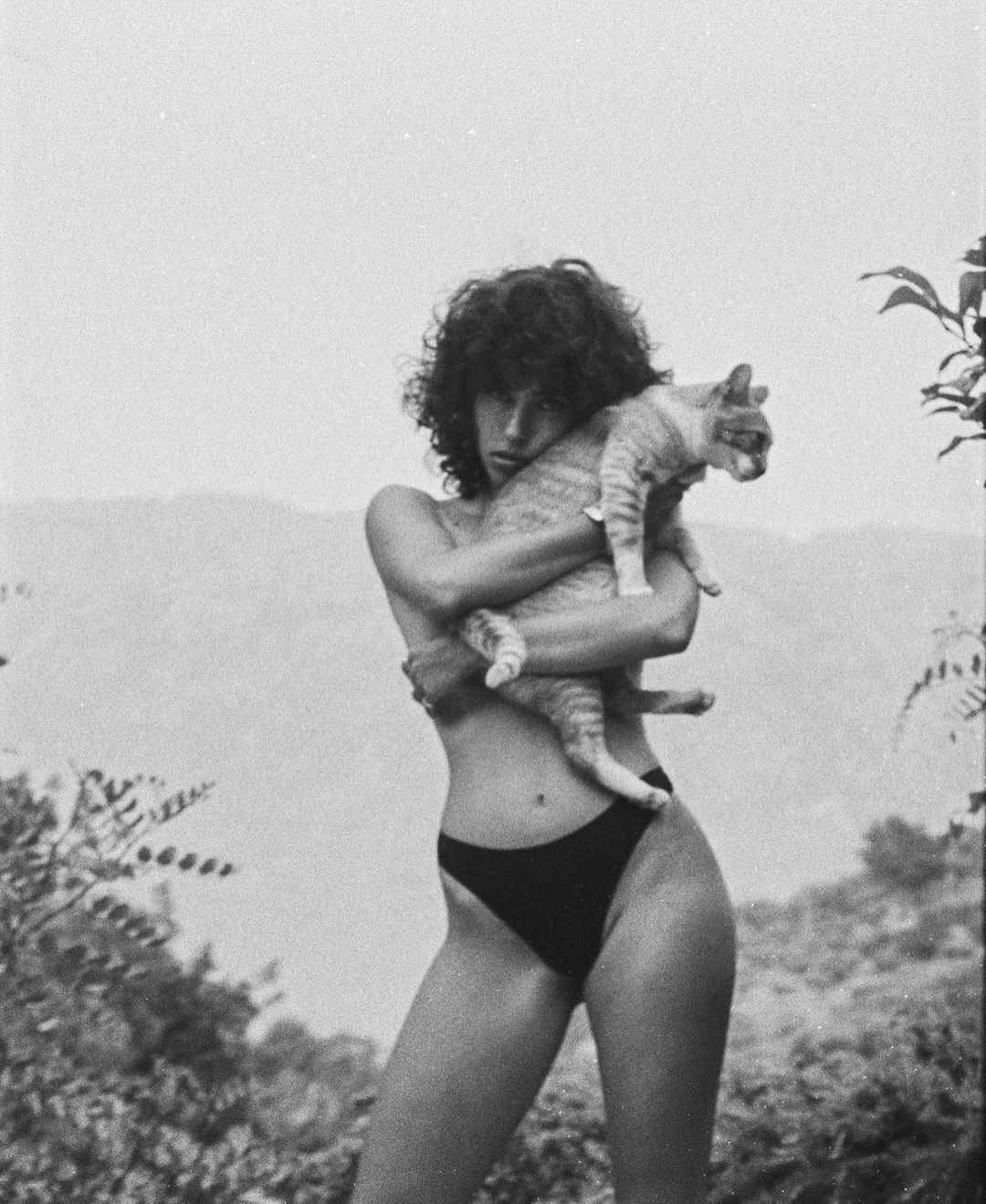 Monochrome portrait of a slender young adult with curly hair holding a striped tabby cat against a natural, hilly background.
