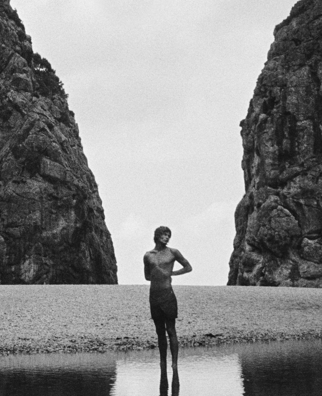 Monochromatic image of a slender male figure standing in shallow water near a beach, with large rock formations in the background.