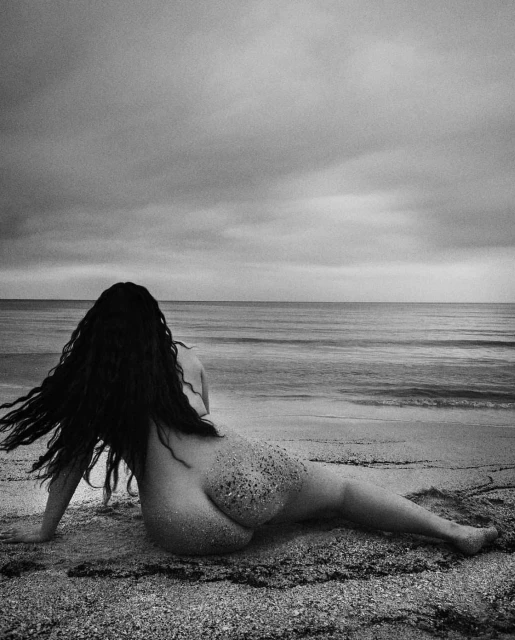 Monochrome photo of a female figure reclining on a sandy beach, looking out at the ocean under a cloudy sky, exuding a serene atmosphere.