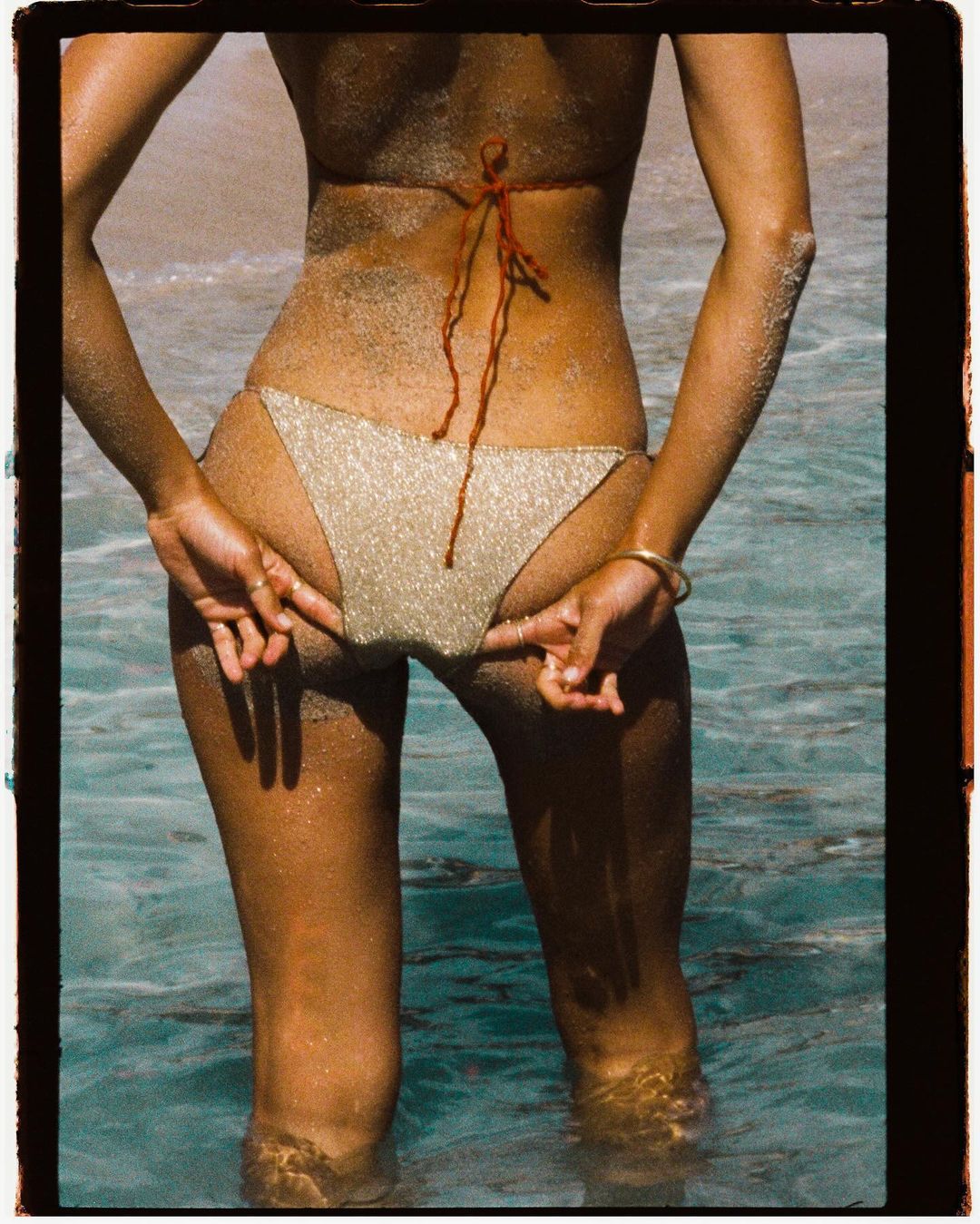 Close-up rear view of a woman wearing a glittering bikini standing in shallow ocean water with sandy beach visible behind her.