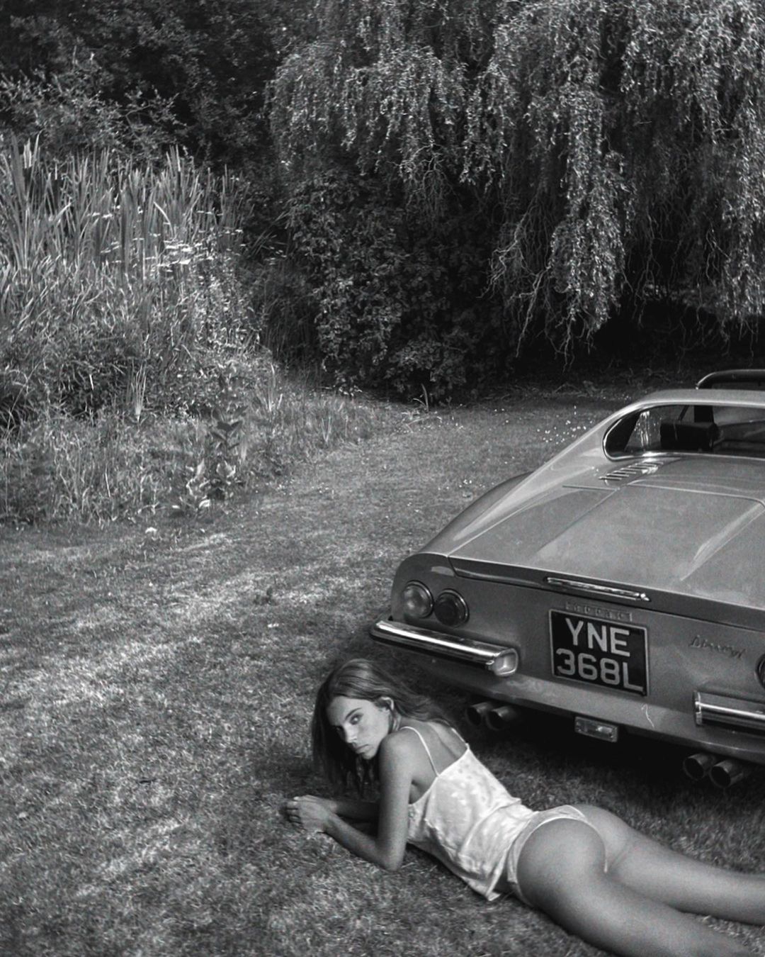 Woman poses beside a vintage car on grassy ground, filtered black and white. She is lying down looking directly at the camera.