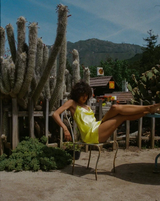 A young woman with curly hair in a yellow dress sits on a metal chair outdoors amidst cacti, set against a backdrop of mountains and sky.
