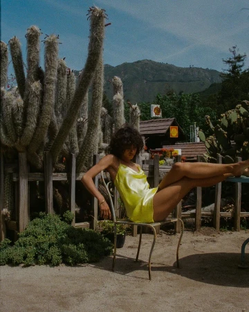 A young woman with curly hair in a yellow dress sits on a metal chair outdoors amidst cacti, set against a backdrop of mountains and sky.