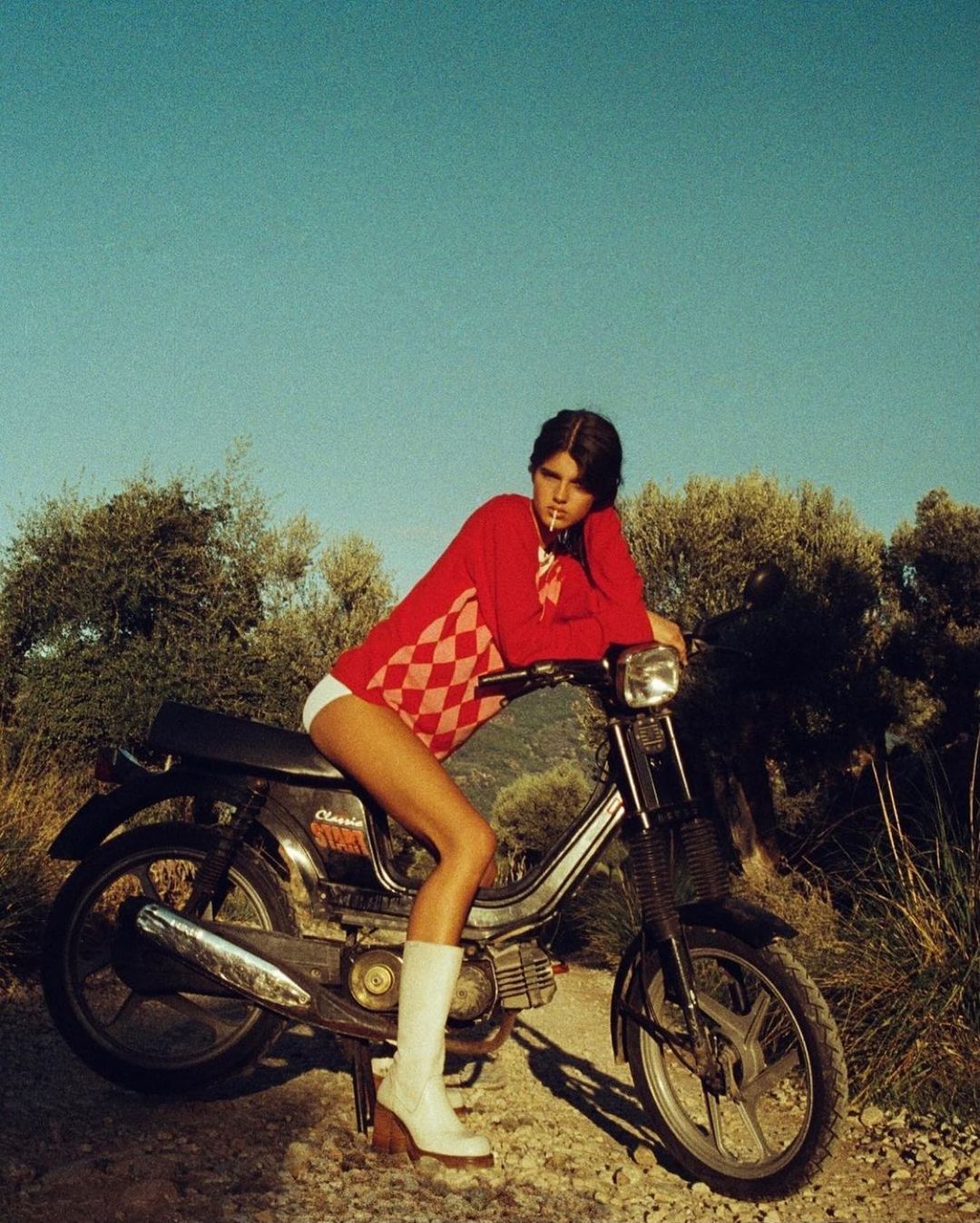 Young woman in red sweater sitting on a vintage moped with a blue sky and grassy trees in the background.