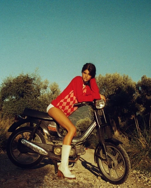 Young woman in red sweater sitting on a vintage moped with a blue sky and grassy trees in the background.