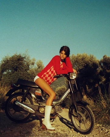 Young woman in red sweater sitting on a vintage moped with a blue sky and grassy trees in the background.