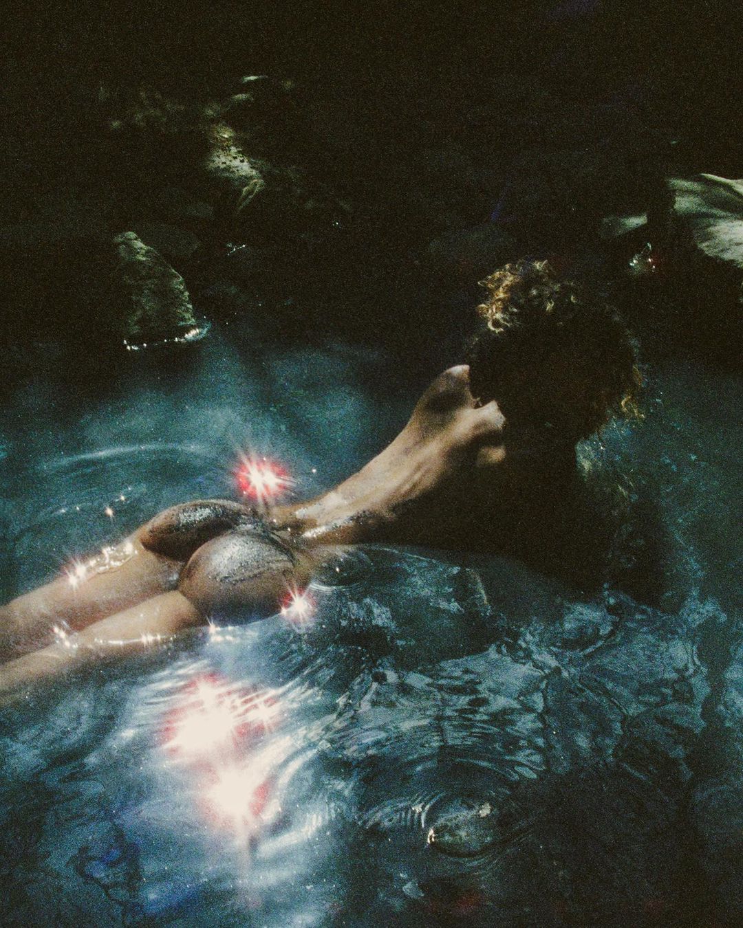 Woman swims in dark water with rocky surroundings and sparkling sunlight reflecting on the water's surface, creating a serene atmosphere.