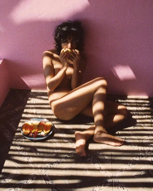 Person seated, eating fruit indoors with shadows creating pattern on floor, against purple wall; plate of fruit, relaxed atmosphere.