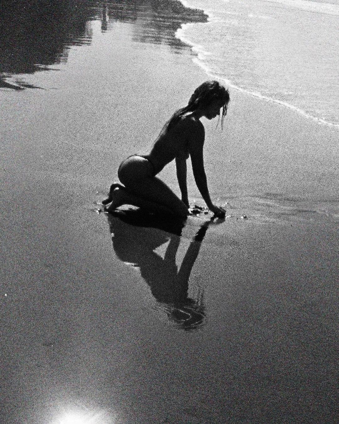 Monochrome image of a woman kneeling on a sandy beach near the water's edge, casting a reflection in the wet sand.