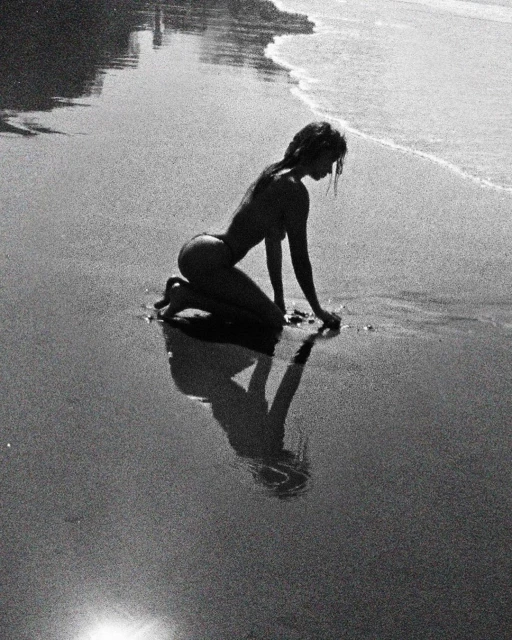 Monochrome image of a woman kneeling on a sandy beach near the water's edge, casting a reflection in the wet sand.