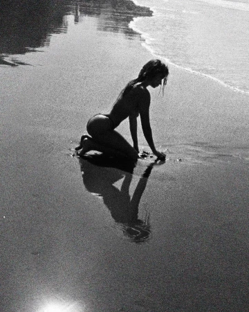 Monochrome image of a woman kneeling on a sandy beach near the water's edge, casting a reflection in the wet sand.