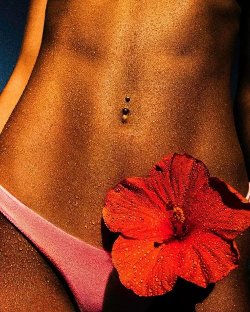 Close-up of a woman's tanned midsection with a navel piercing, wearing a pink bikini bottom, and adorned with a red hibiscus flower.