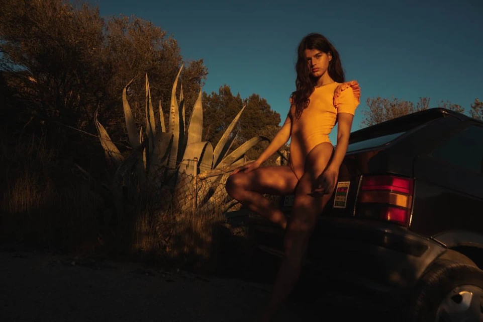 Adult female model poses on a car in the desert with cacti, bathed in warm sunset light and captured in a medium shot.