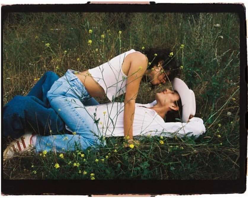 Two people lying together in a grassy meadow surrounded by wild vegetation, captured in a horizontal analogue frame.