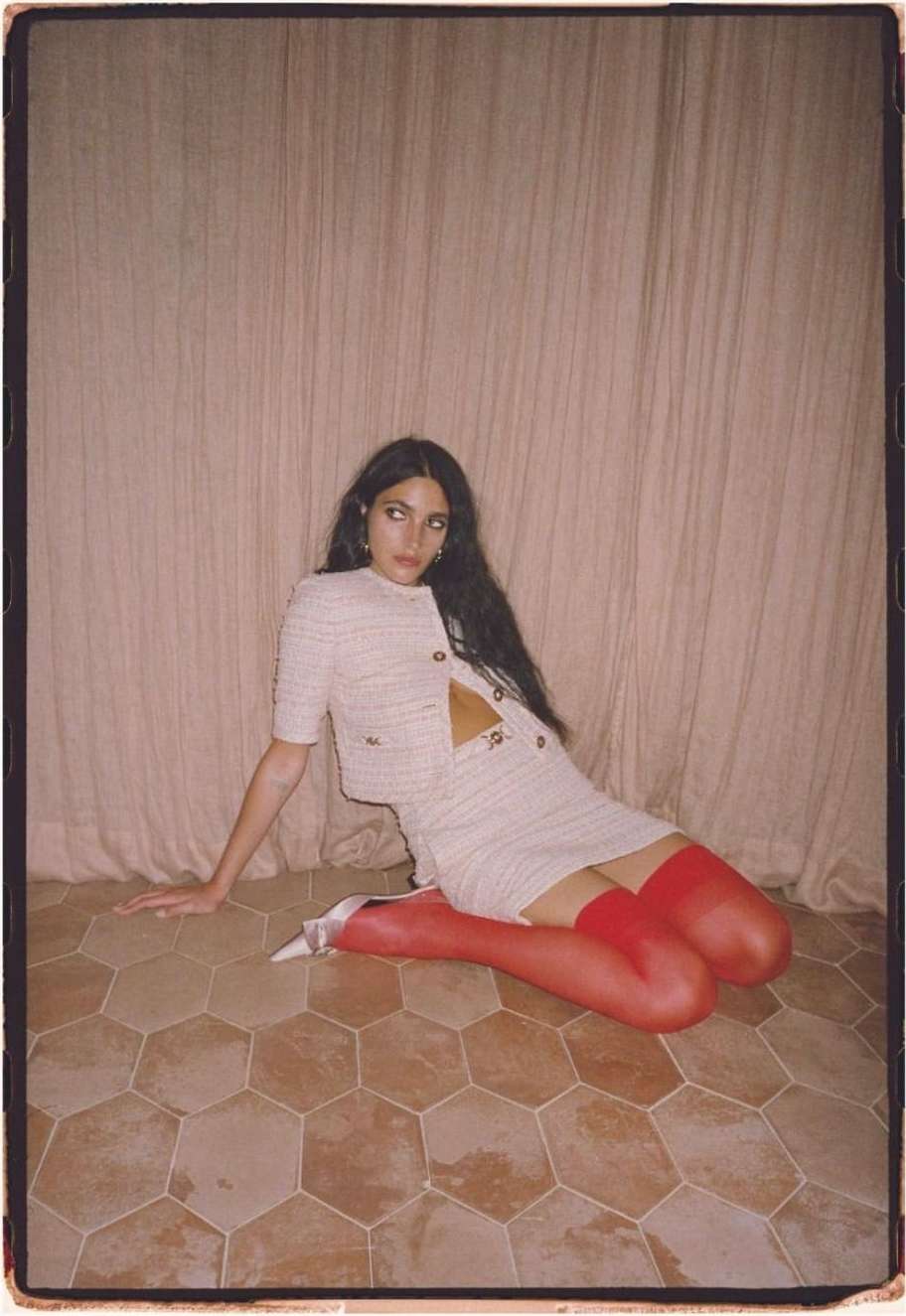 A young woman with dark hair sits in a skirt and jacket, paired with red stockings and white heels, posing in front of a neutral-toned curtain.