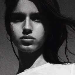 Close-up monochrome portrait of a young woman with dark hair blowing in the wind, wearing a white t-shirt and hoop earrings.