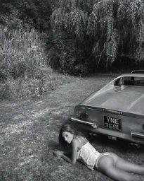 Woman lying on grass next to the rear of a parked sports car under lush trees in a black and white photograph.