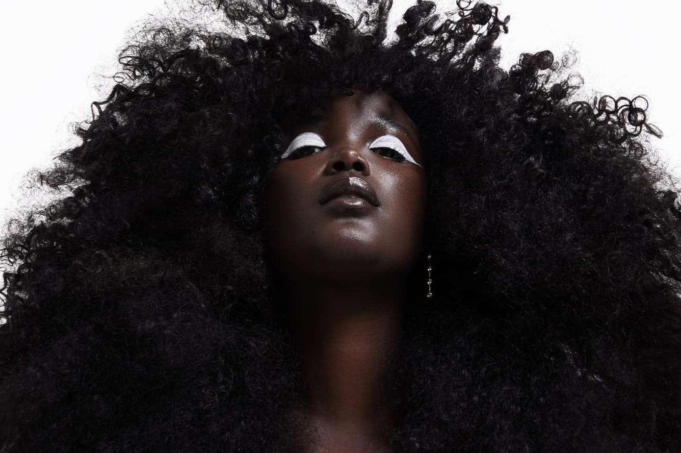 Portrait of a woman with dark skin and an afro looking upward, wearing white makeup around her eyes, set against a plain white backdrop.