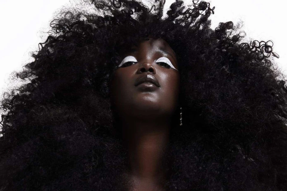 Portrait of a woman with dark skin and an afro looking upward, wearing white makeup around her eyes, set against a plain white backdrop.