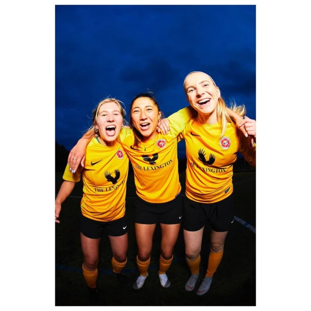 Three smiling women in yellow soccer jerseys embrace on a green field under a blue sky, celebrating victory with visible joy and camaraderie.