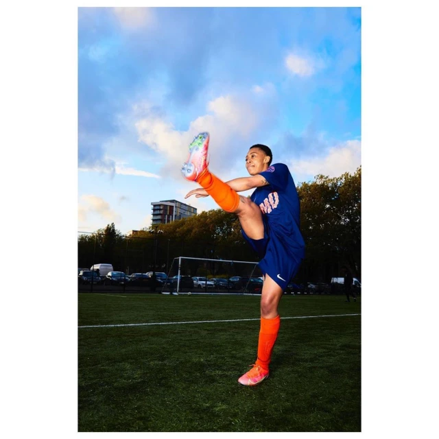Young woman in soccer uniform dynamically kicks a soccer ball on a green field under a partly cloudy sky in an action shot.