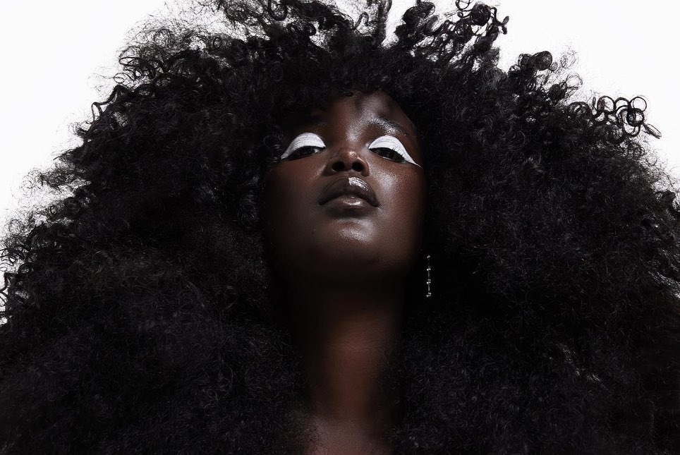 Glamour portrait of a Black woman with striking white eye makeup, lush afro hair, and shimmering lips against a stark white backdrop.