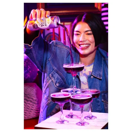 Smiling woman pouring sparkling drink from a can into a tower of coupe glasses on a white marble surface against a dark background.