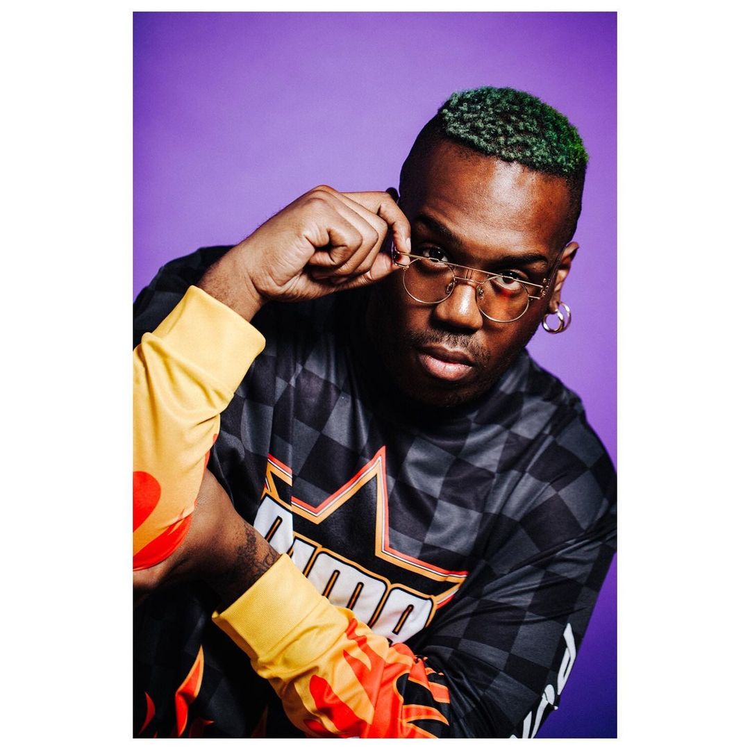 Stylish man in Puma jacket and glasses poses against a purple background, showcasing a modern, casual fashion statement.