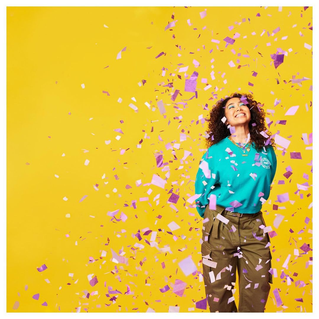 A happy adult female stands smiling on a yellow background while purple confetti falls, looking up in a medium shot, casual fashion.