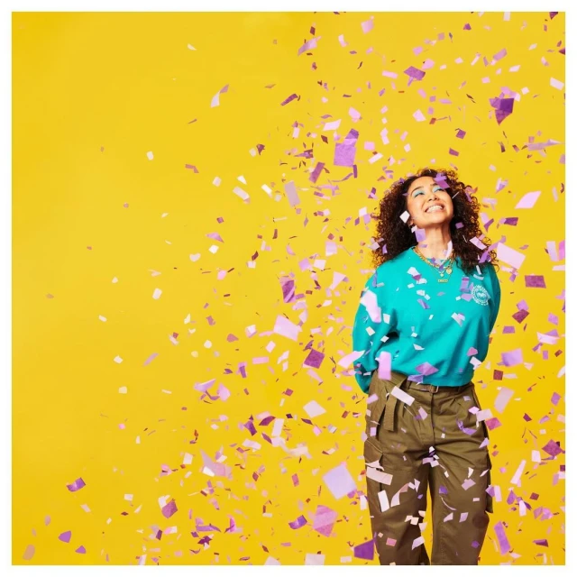 A happy adult female stands smiling on a yellow background while purple confetti falls, looking up in a medium shot, casual fashion.