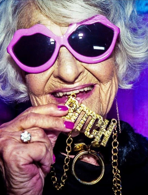 Elderly woman with white hair wears pink lip-shaped sunglasses and holds a gold necklace with the word "BITCH" in rhinestones.