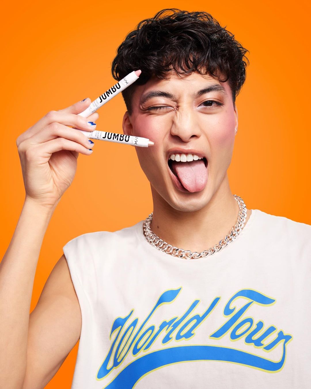 Young Asian man winking, tongue out, in a sleeveless shirt with 'World Tour' and chunky chain, holding two Jumbo makeup pencils.