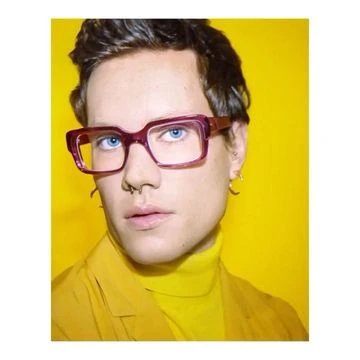 Close-up studio shot of a person with blue eyes and glasses, wearing a yellow sweater, set against a bright yellow background.