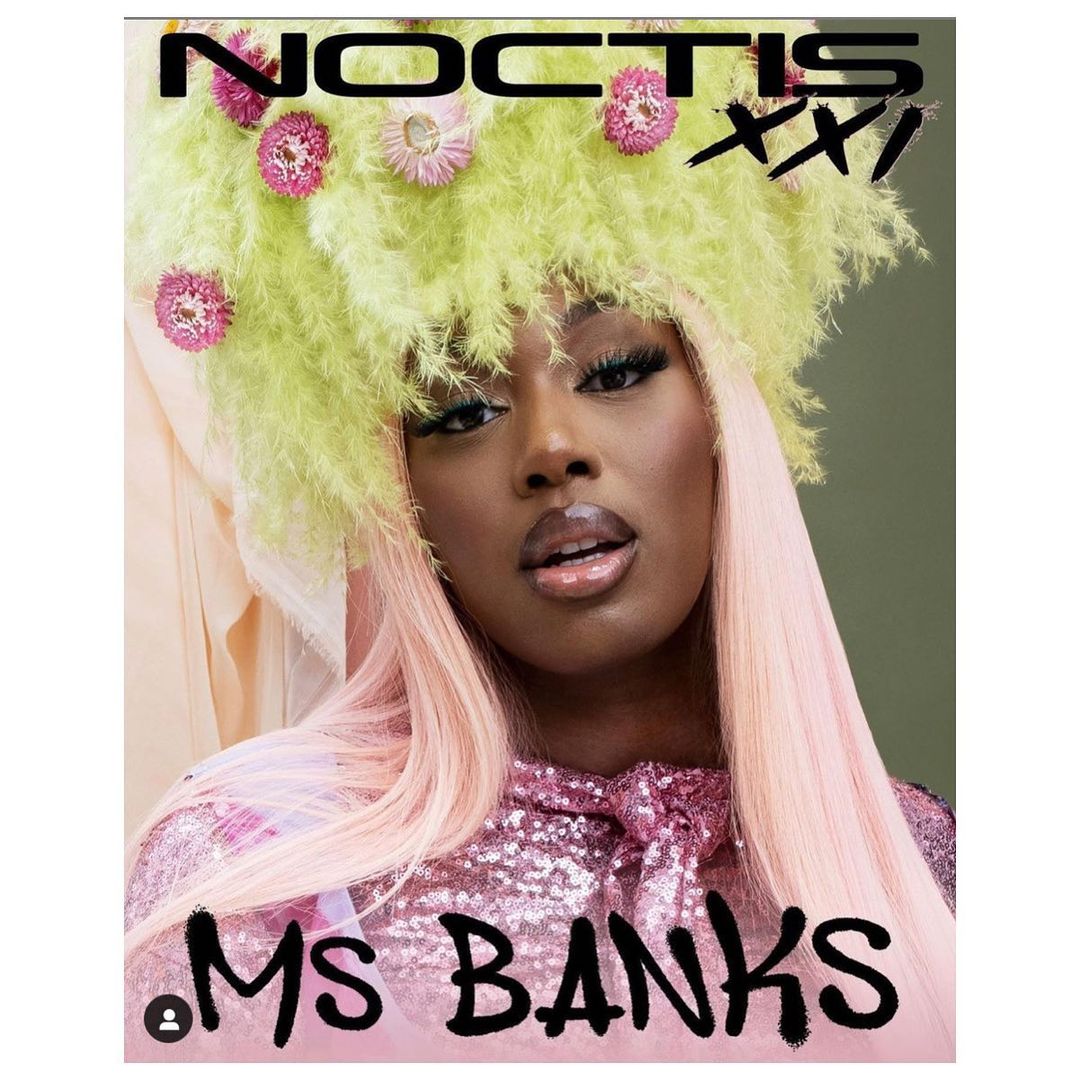 Dark-skinned woman with decorative headpiece and pink wig poses in a shiny pink top against a green and peach backdrop. 'Ms Banks' text.