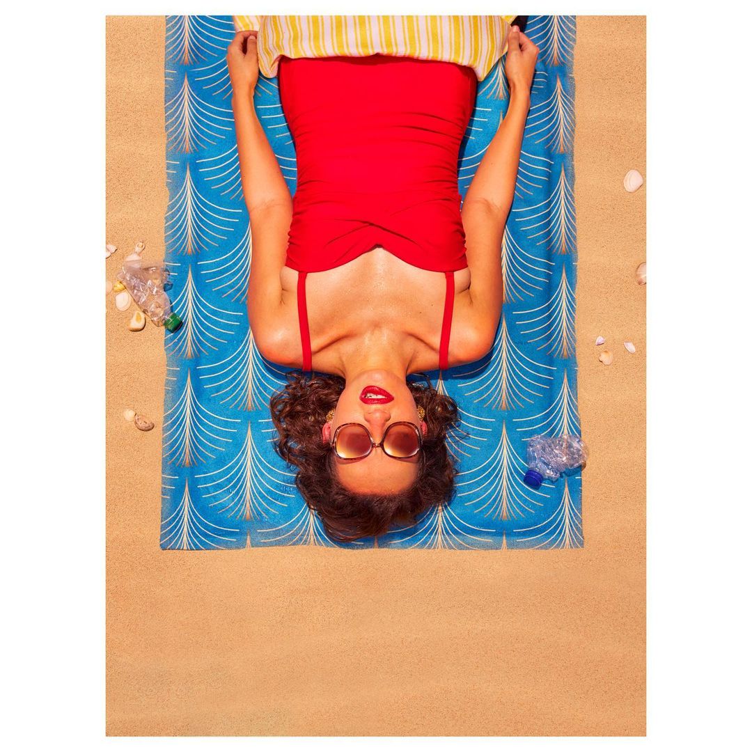 Overhead view of a woman in a red swimsuit lying on a patterned beach towel on sand, with plastic bottles and shells around her.