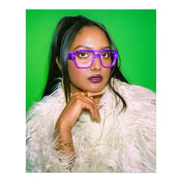 Close-up studio shot of an adult female with dark hair, wearing purple eyeglasses, gold jewelry, and a white faux fur coat.