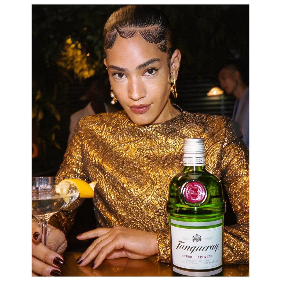 Elegant adult female in golden dress poses at a bar with a Tanqueray gin bottle and martini glass in a dimly lit indoor setting.
