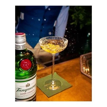 A green Tanqueray gin bottle and a stemmed cocktail glass with liquid on a dark brown surface with a green cloth napkin.