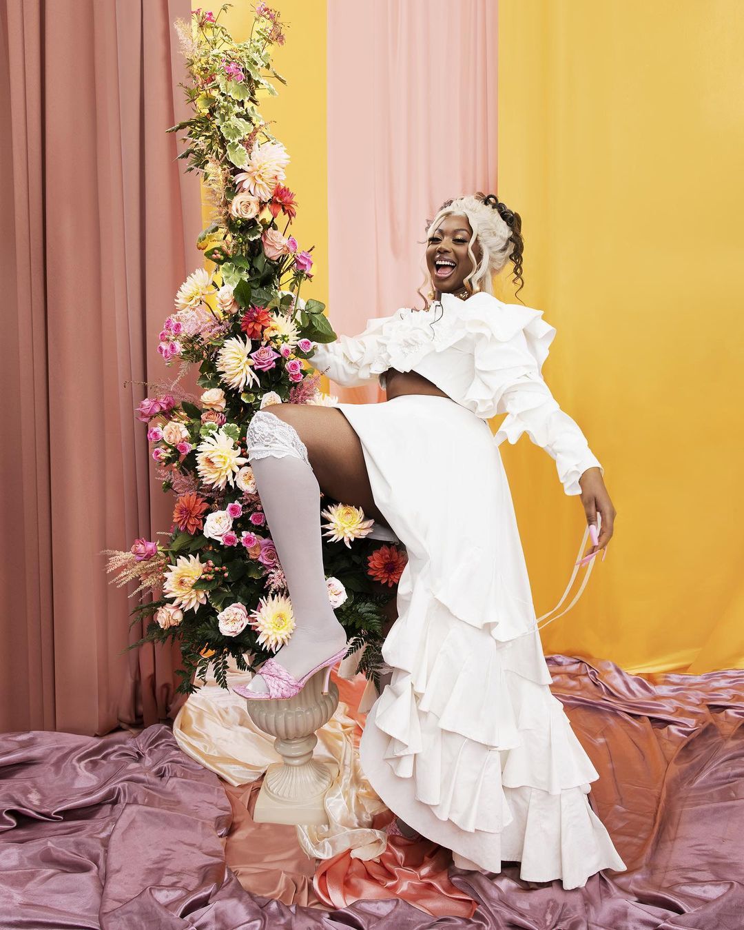 Woman in white outfit poses with leg raised, wearing white stockings and pink heels, next to a large flower arrangement in a studio setting.