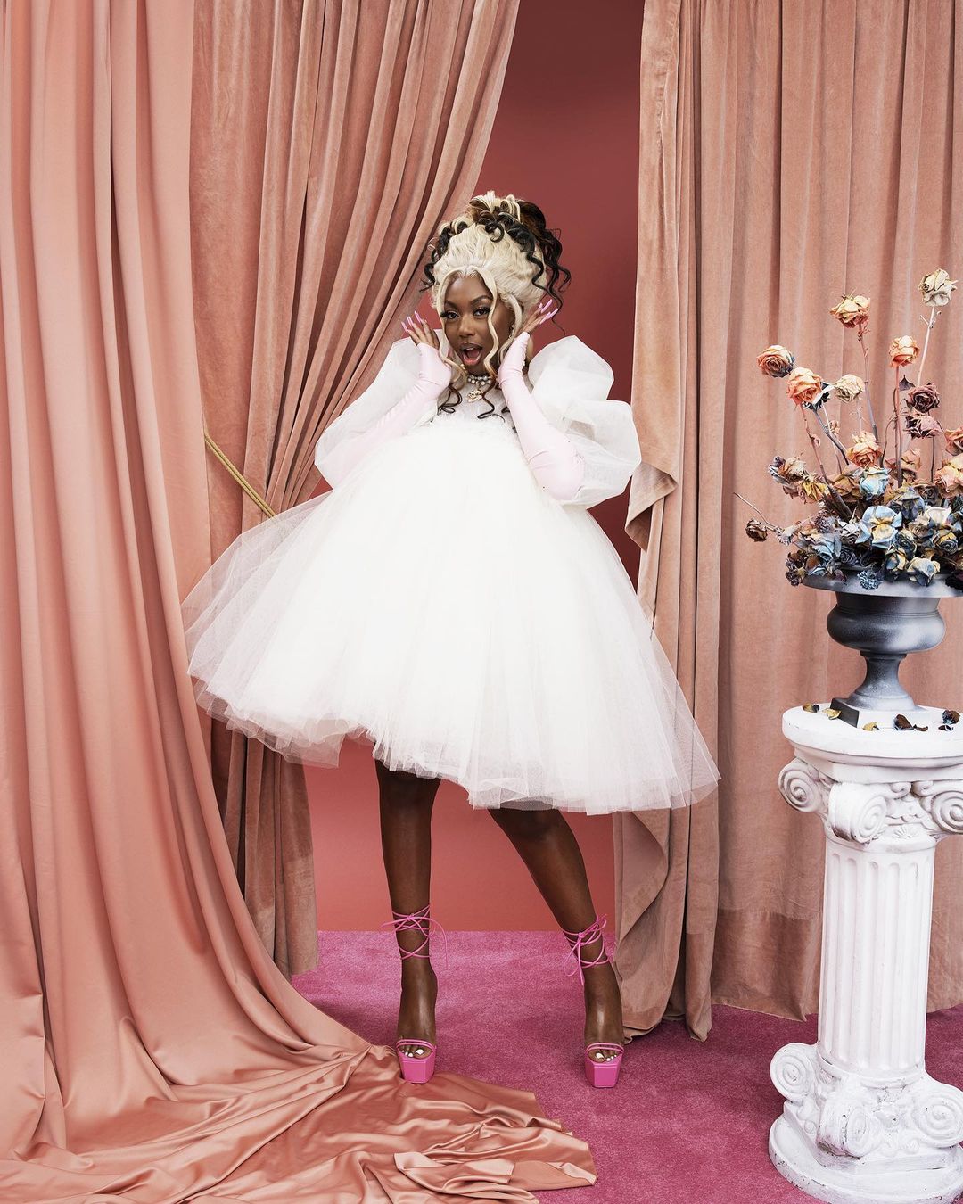 Elegant woman in a white tulle dress and pink heels poses amid pink curtains and a column adorned with floral arrangements in an indoor setting.