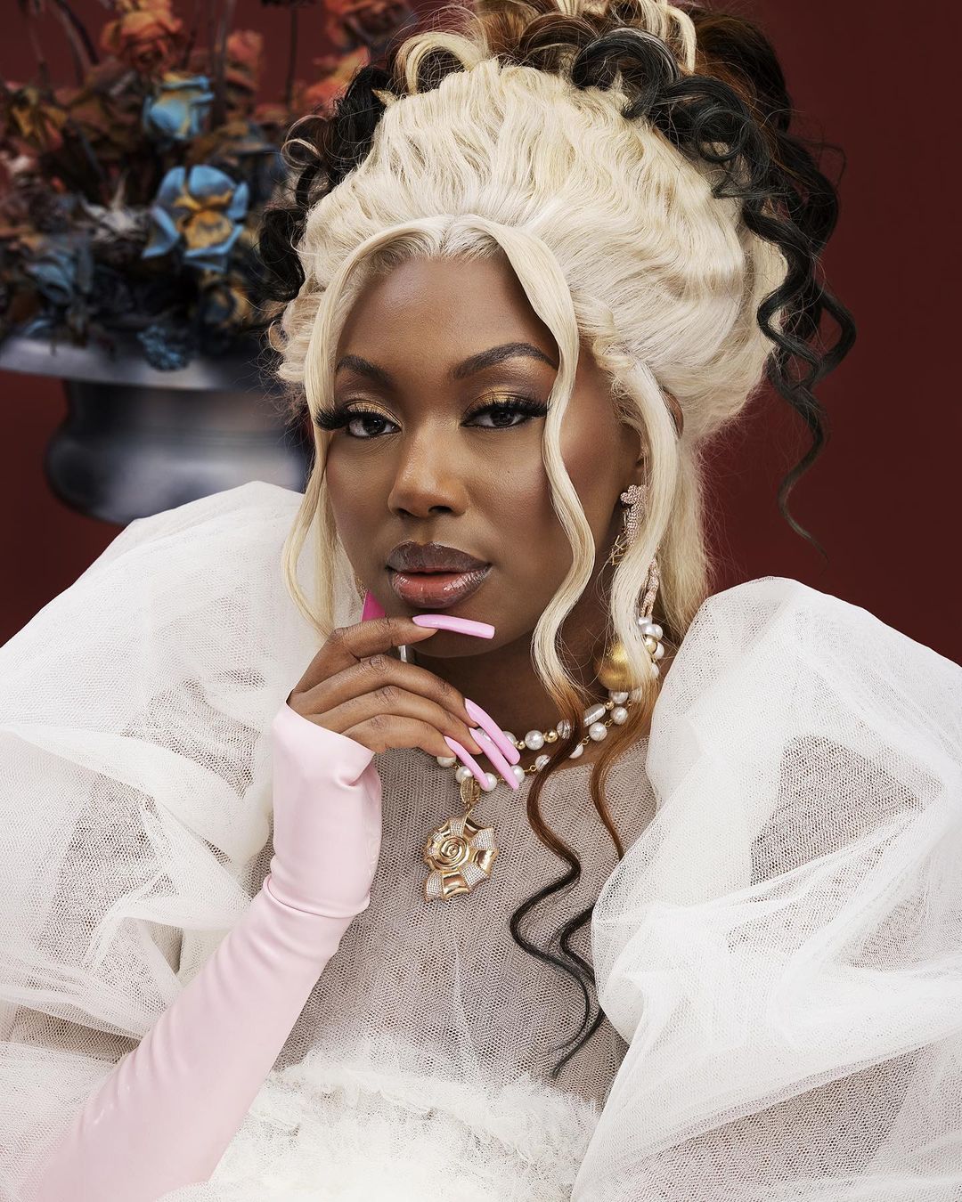 Elegant portrait of a woman with dramatic makeup and hairstyle, wearing a white tulle gown, set against a dark red background.