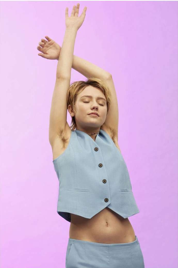 Woman in a light blue vest and pants poses with her arms raised on a pastel purple background in a studio-lit, medium shot.