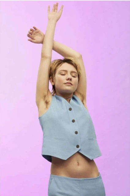 Woman in a light blue vest and pants poses with her arms raised on a pastel purple background in a studio-lit, medium shot.
